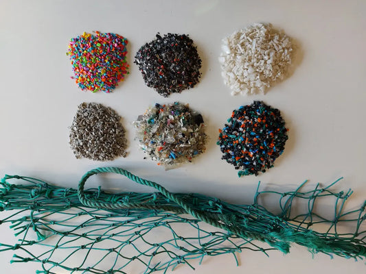 Six piles of recycled ocean plastic granulate in various colours alongside a green fishing net